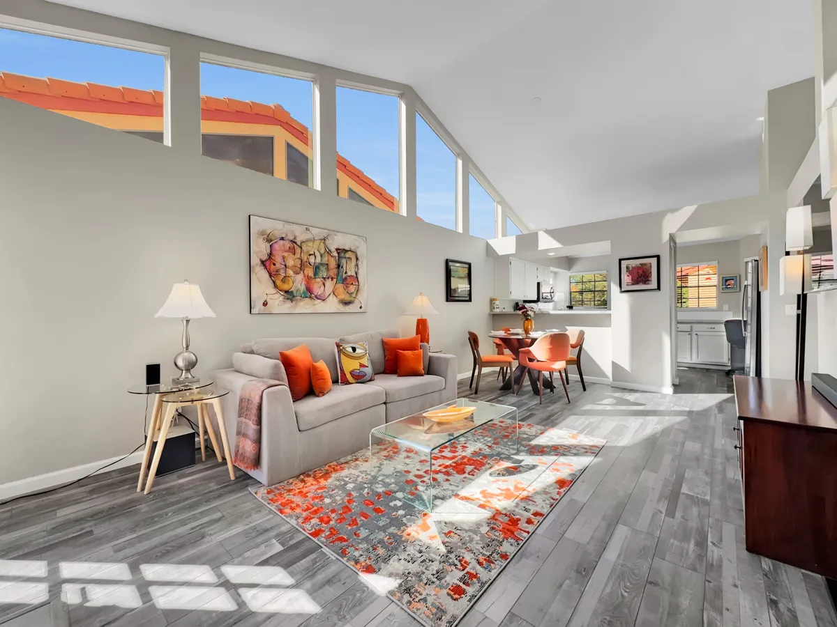 A cozy contemporary living room with a sofa adorned with colorful cushions, a glass coffee table, eye-catching artwork on the wall, and a dining area visible in the background. The space feels bright and inviting, ideal for relaxing or entertaining guests.