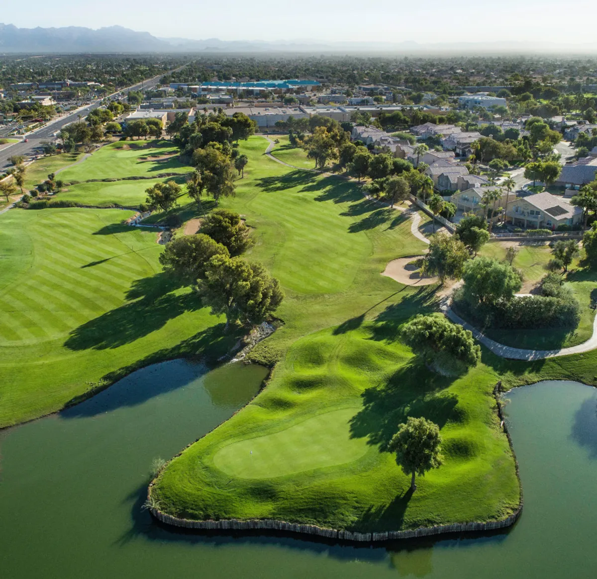 A scenic view of a lush green park with a tranquil lake in the foreground and a well-maintained golf course. Residential buildings are visible in the background, and the setting sun casts a warm glow over the landscape, creating an inviting and relaxed atmosphere.