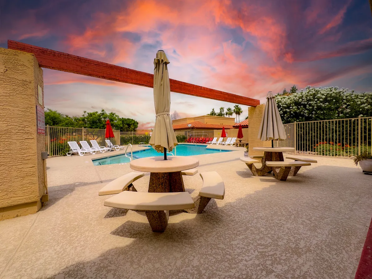 A vibrant pool area with picnic tables, umbrellas, and lush landscaping, set against the soft light of sunset. People relax by the water, socializing and enjoying the outdoor spaces within the community.