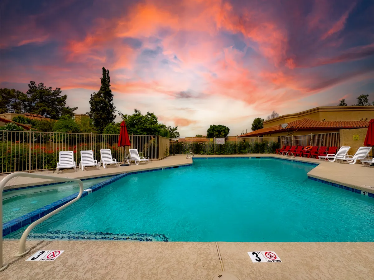 A serene community swimming pool is surrounded by neatly landscaped gardens, with comfortable lounge chairs and vibrant red umbrellas set against a beautiful sunset sky. The pool area is clearly designed for both relaxation and social gatherings, evoking a resort-like atmosphere ideal for unwinding after work.