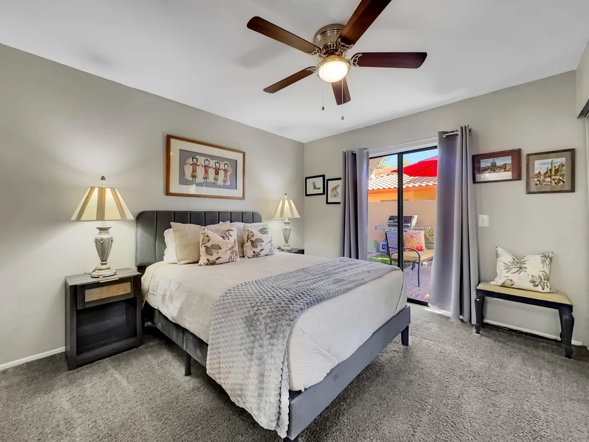 A tranquil bedroom setup with a bed adorned with decorative pillows, flanked by two bedside lamps and framed artwork. A sliding glass door opens to the outdoors, suggesting immediate access to a private yard or patio.