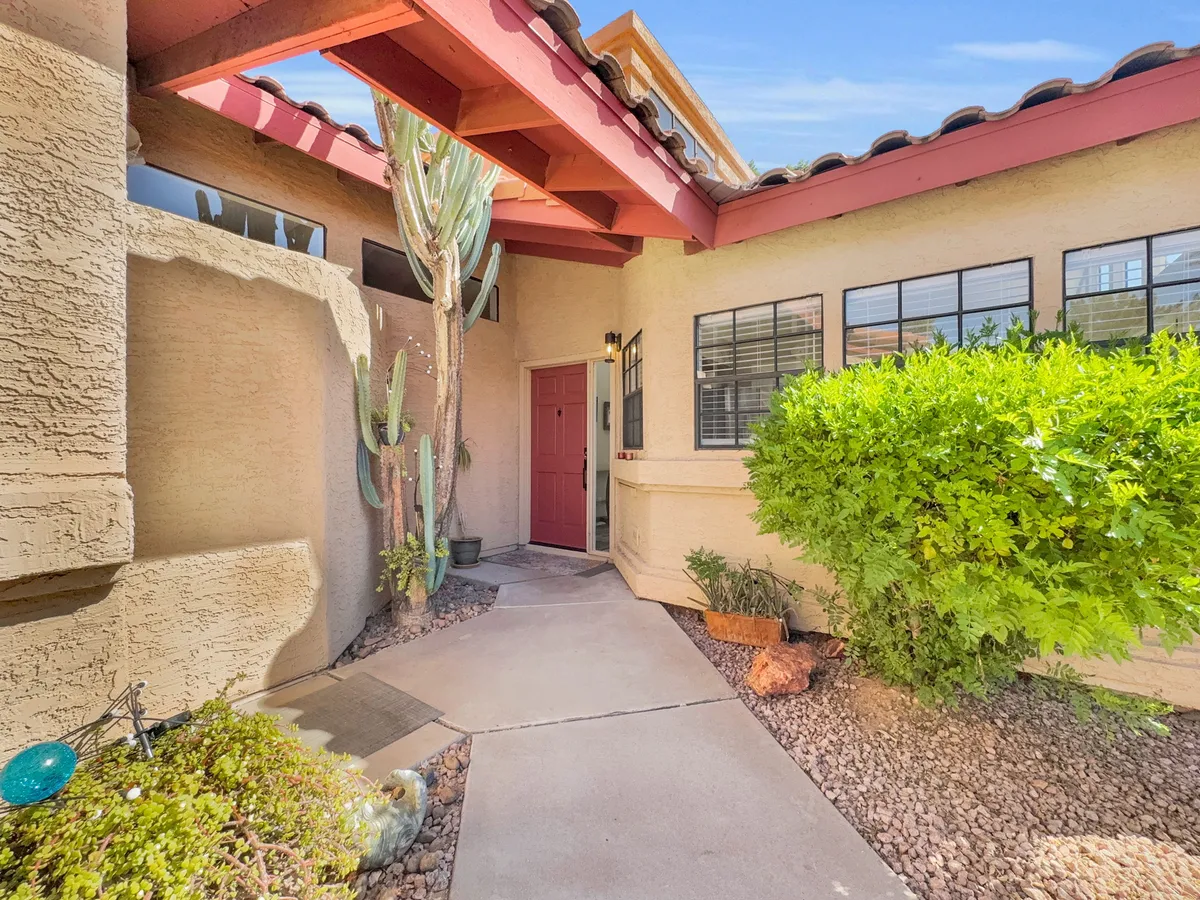 A welcoming entrance framed by a striking red door, surrounded by native cacti and lush green shrubs, with textured walls adding Southwestern charm. The scene hints at both arrival and the inviting landscape that defines Arizona living.