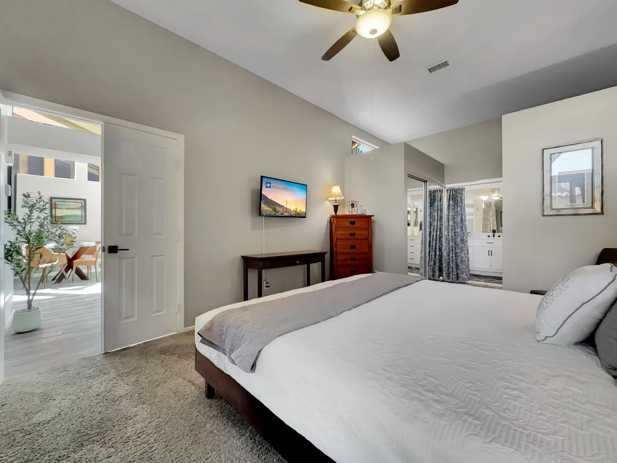 A serene bedroom featuring a light-colored bed, mounted TV, and a nightstand with a lamp. The open door leads directly to an outdoor area, suggesting seamless indoor-outdoor living and inviting natural light into the room.