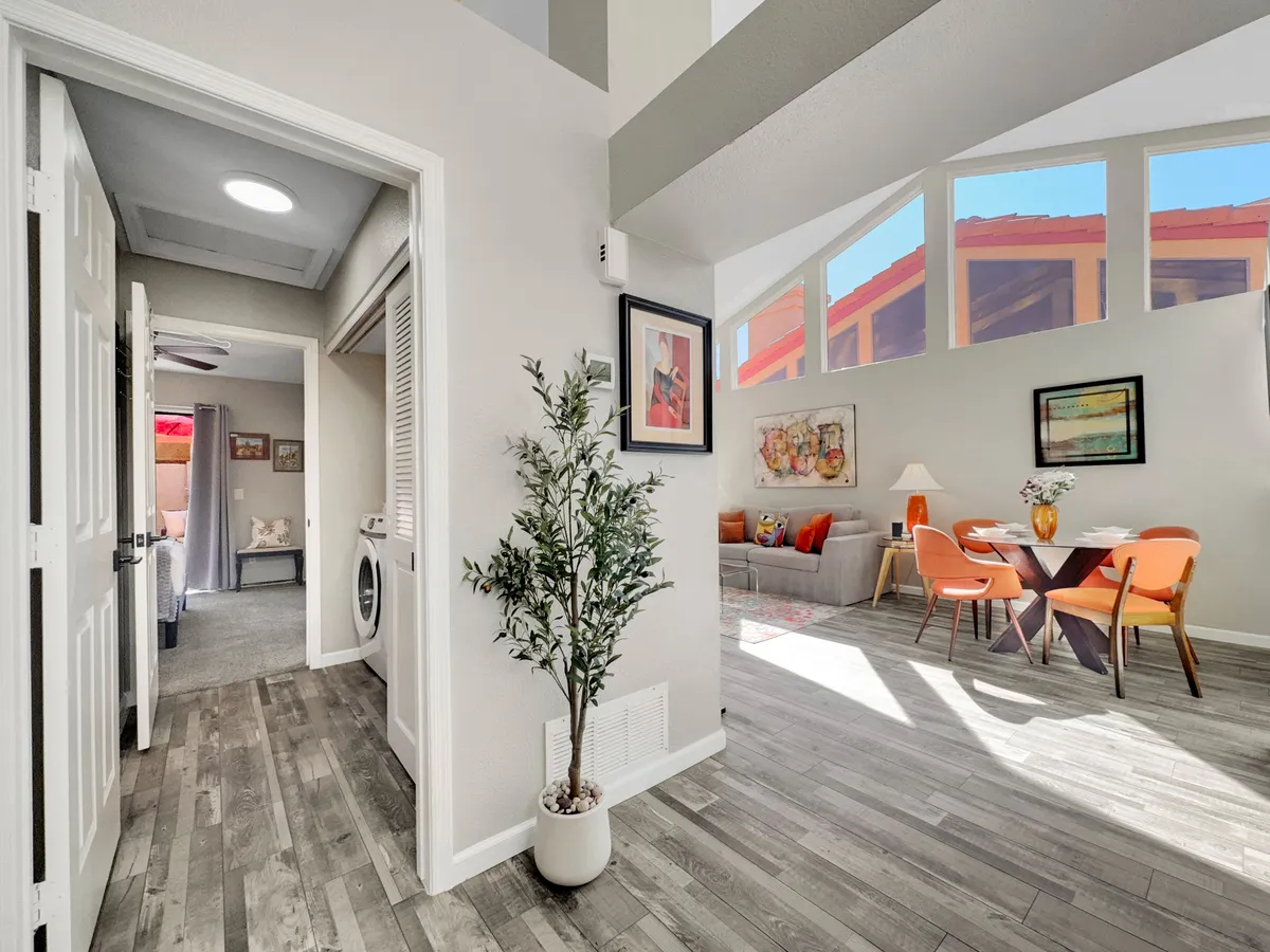This image depicts an open-plan dining area with a modern table surrounded by vibrant orange chairs, colorful artwork adorning the wall, a lush indoor plant, and high ceilings that add to the sense of spaciousness. The living room area is visible, offering an inviting glimpse of the plush seating beyond.