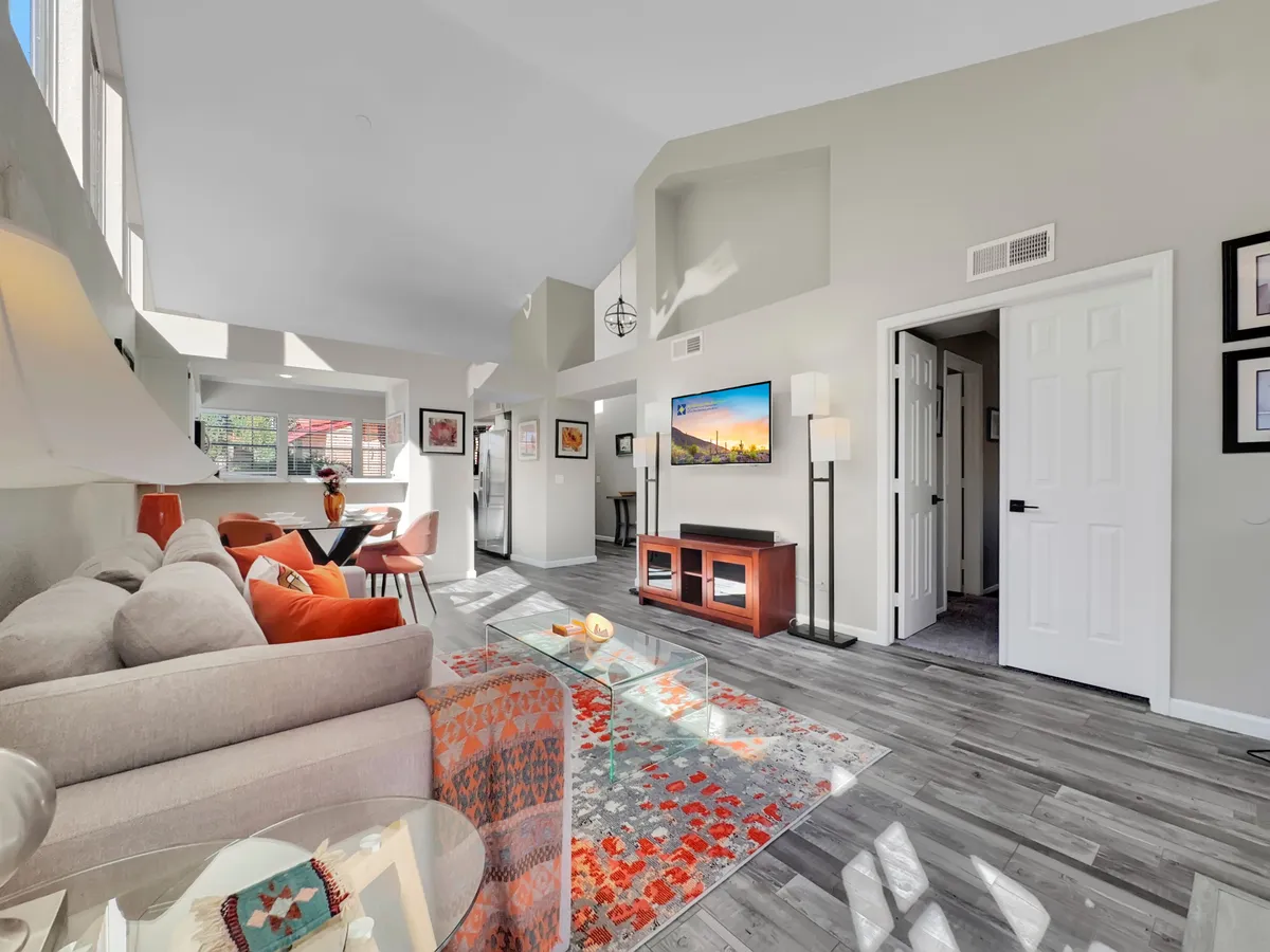 A sunlit living room featuring a sofa with colorful cushions, a sleek coffee table, large windows letting in abundant natural light, a TV mounted on the wall, and modern artwork adding character. The room exudes warmth and is well-suited for both downtime and entertainment.