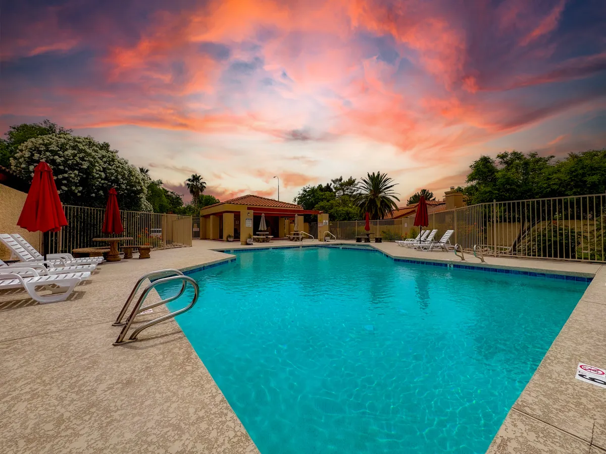 This scene showcases a sparkling community swimming pool surrounded by red umbrellas, lounge chairs, and lush landscaping. The pool is well-maintained and exudes a relaxed, cheerful resort vibe, inviting guests to lounge, swim, or socialize. The late afternoon lighting adds warmth and a touch of Arizona luxury.