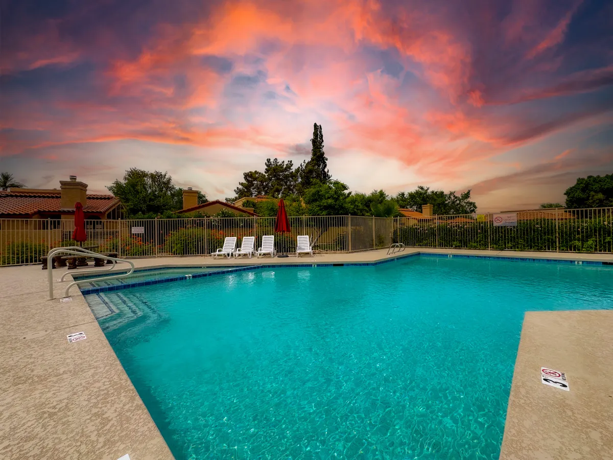 A serene outdoor swimming pool scene with inviting lounge chairs and bold red umbrellas set beneath a colorful Arizona sky. The pool area is designed for relaxation and social gatherings, offering ample space to sunbathe or enjoy drinks poolside while taking in the sunset.